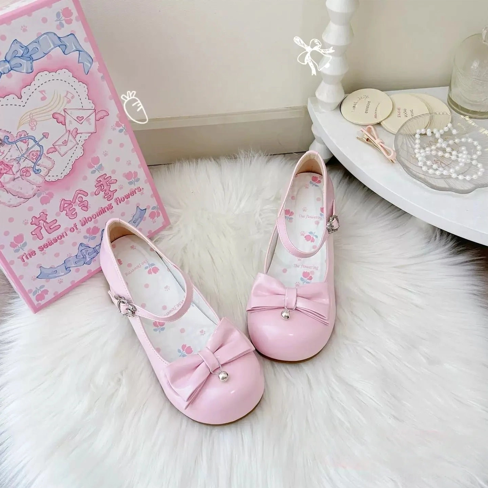 Pink glossy patent leather sweet lolita Mary Jane shoes with pearl-adorned front bow, adjustable ankle buckle strap, floral printed inner lining, and chunky low block heel, displayed on white textured fur rug with themed gift box packaging and vanity accessories