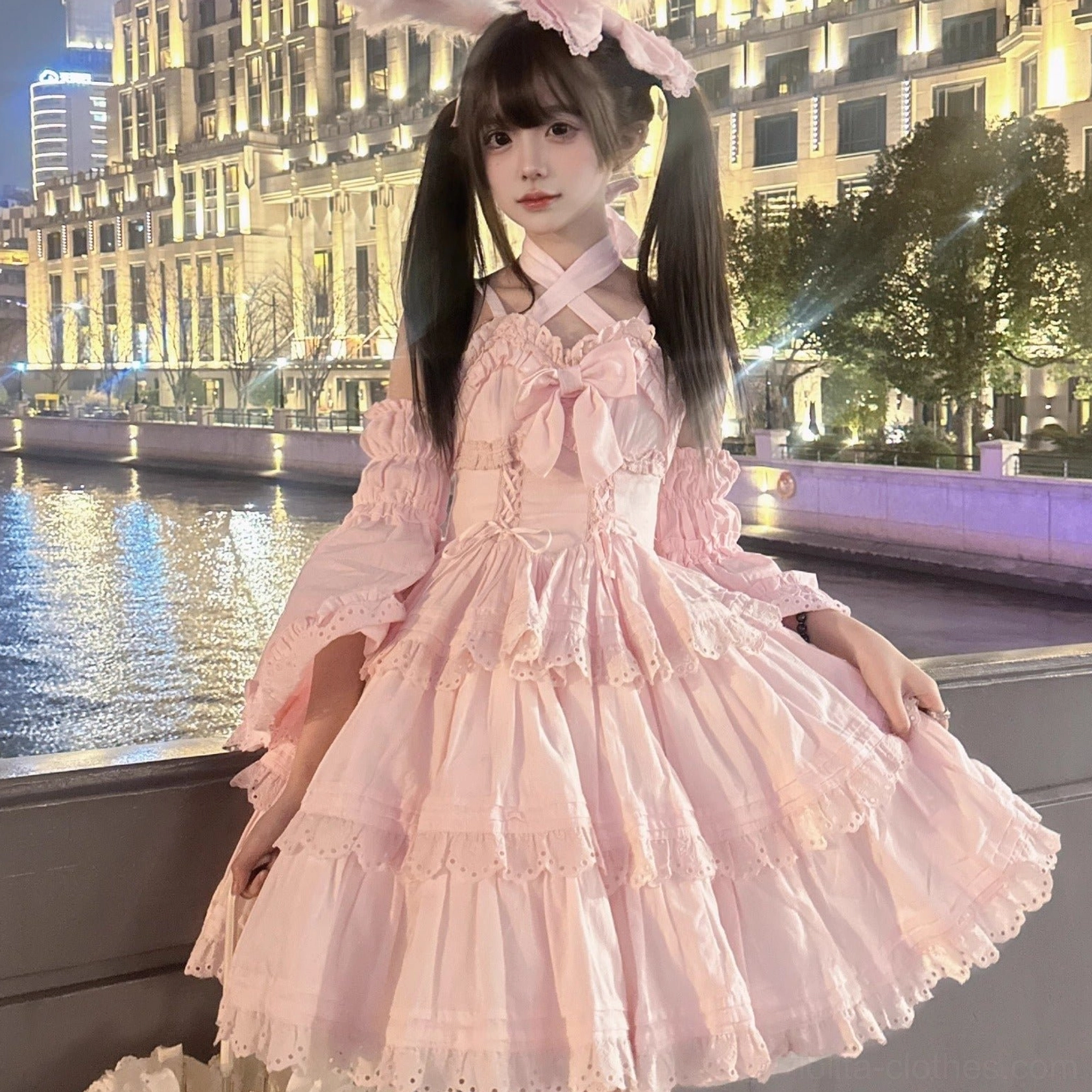 Pink sweet lolita one-piece dress featuring cross halterneck design with front bow, lace-trimmed tiered skirt, detachable puff sleeves, worn by model at twilight riverside urban setting