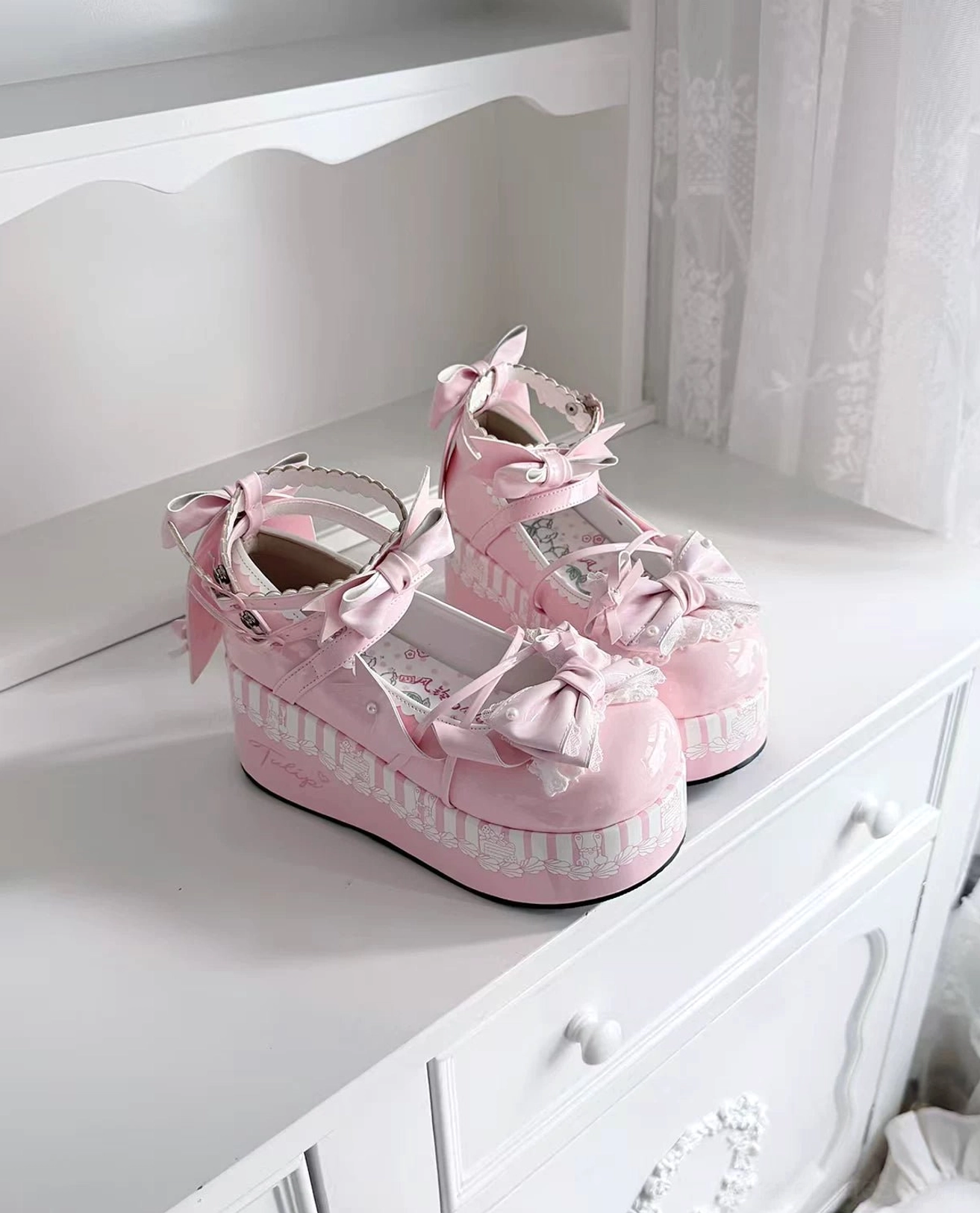 Pink sweet lolita platform shoes with ankle strap bow, striped platform midsole, lace trimmed upper held in hand showing two pairs on white vanity table