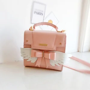 Pink sweet Lolita shoulder bag with white angel wings and pink bow decoration, displayed on indoor Paris-themed backdrop showing matte finish and gold hardware details