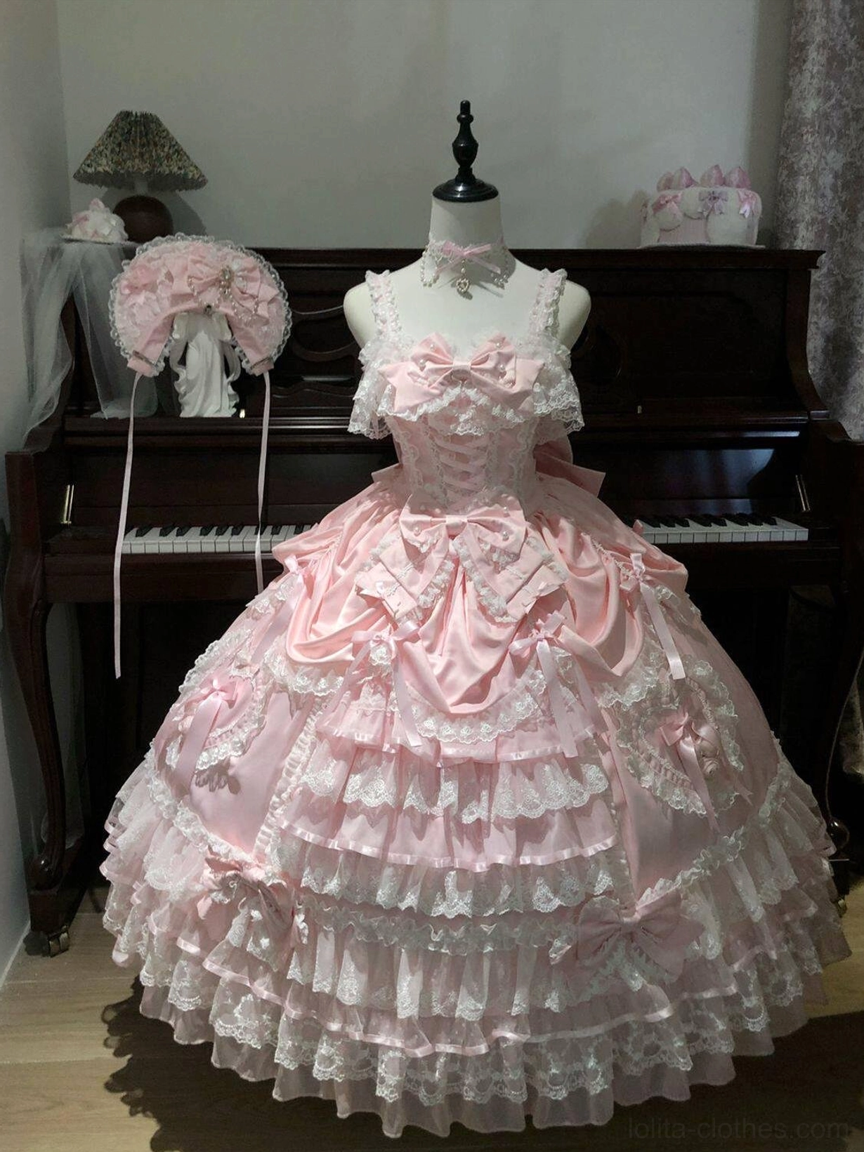 Pink sweet wedding Lolita JSK dress on dress form indoor studio display with sweetheart neckline, satin bodice with bow accents, layered lace tiered skirt, matching pink bonnet beside dress