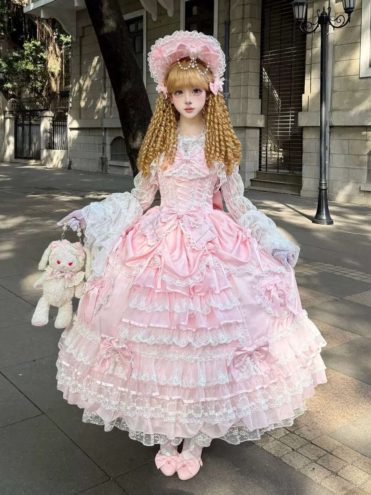 Pink sweet wedding Lolita JSK dress with tiered lace ruffle skirt, petal-shaped bodice, and bow accents, worn with pink bonnet and white lace gloves on blonde curly twin tails model in outdoor retro street setting holding plush rabbit