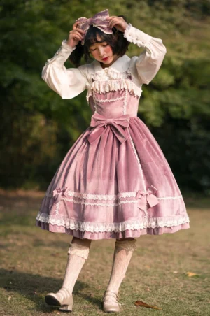 Pink velvet Lolita jumper skirt with white lace trims and layered ruffled hem worn outdoors in natural setting with matching hair bow