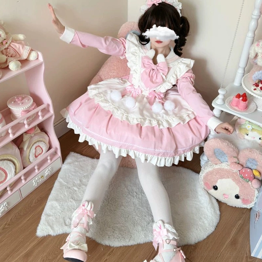 Model wearing pink and white maid lolita OP dress with fluffy pom-pom decorations, ruffled apron and skirt hem, large front chest bow, white tights and pink lace shoes, dynamic standing pose