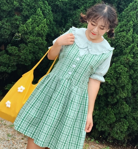 Plus-size model wearing green plaid doll collar lolita short sleeve blouse with puff sleeves and front button placket matched with yellow flower tote bag in outdoor pine forest area showing half-body styling