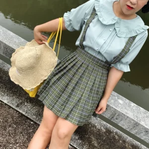 Plus-size model wearing mint green ruffled collar lolita blouse with green plaid pleated suspender skirt and straw tote bag standing on riverside stone platform outdoor garden setting showing complete coordinate