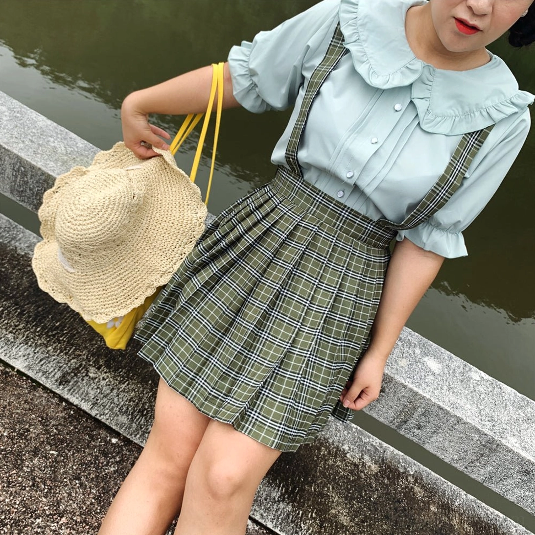 Plus-size model wearing mint green ruffled collar lolita blouse with green plaid pleated suspender skirt and straw tote bag standing on riverside stone platform outdoor garden setting showing complete coordinate