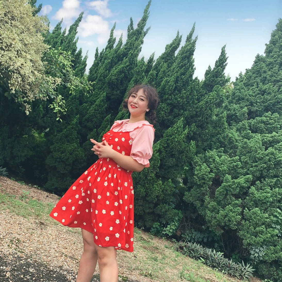 Plus-size model wearing red polka dot suspender dress with pink peter pan doll collar lolita blouse with puff short sleeves standing on outdoor path beside pine forest showing front wearing effect