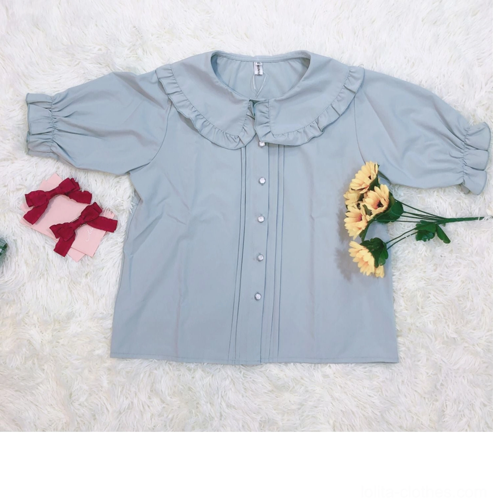 Flat lay top-down view of smoky blue ruffled collar lolita short sleeve blouse with pearl-like front buttons and red bow hair accessories displaying collar detail and craftsmanship on white background