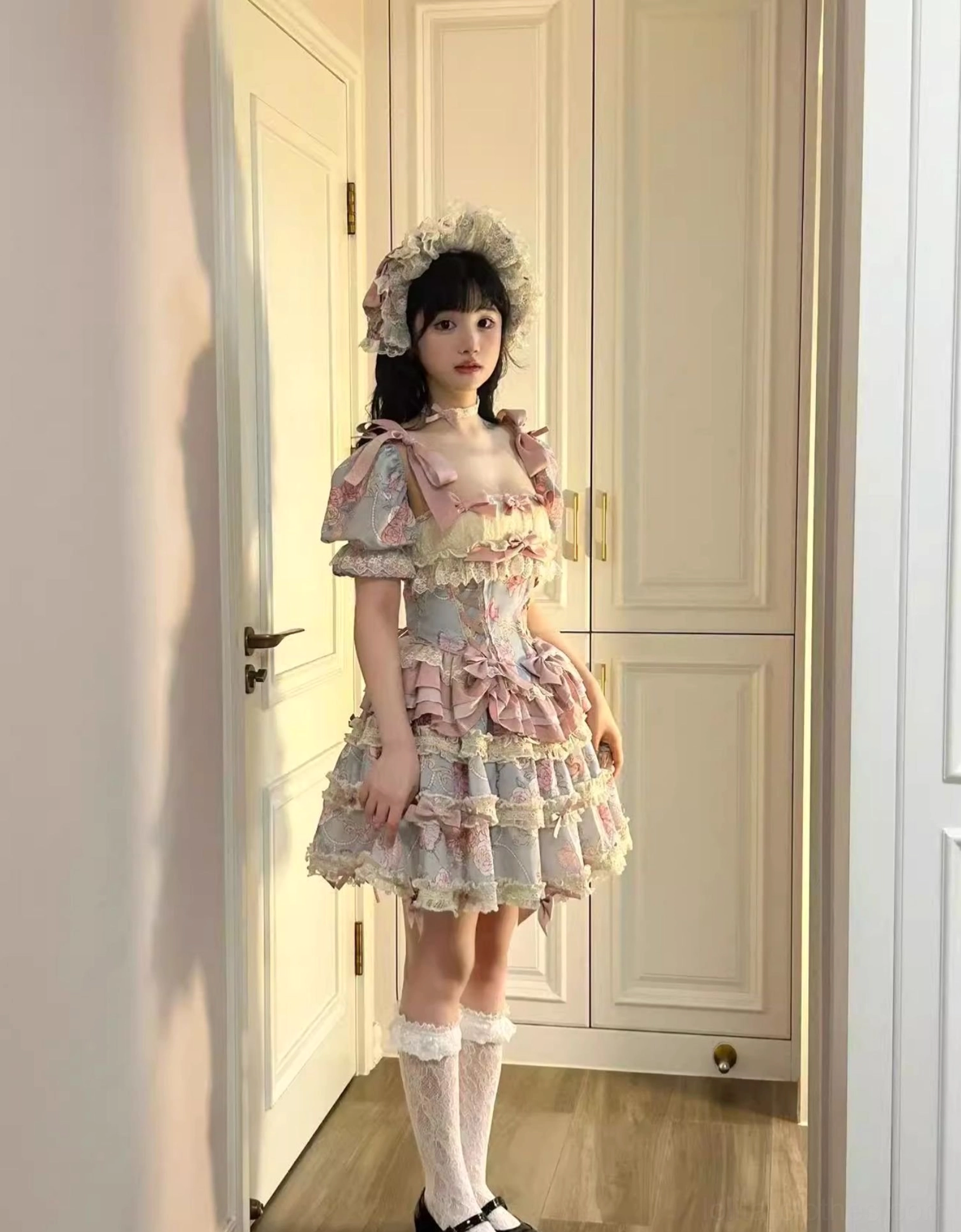 Full-body seated shot of plus size sweet lolita dress set on bedroom bed featuring full multi-layered ruffle skirt cream lace edging pink satin bow decorations on front waist