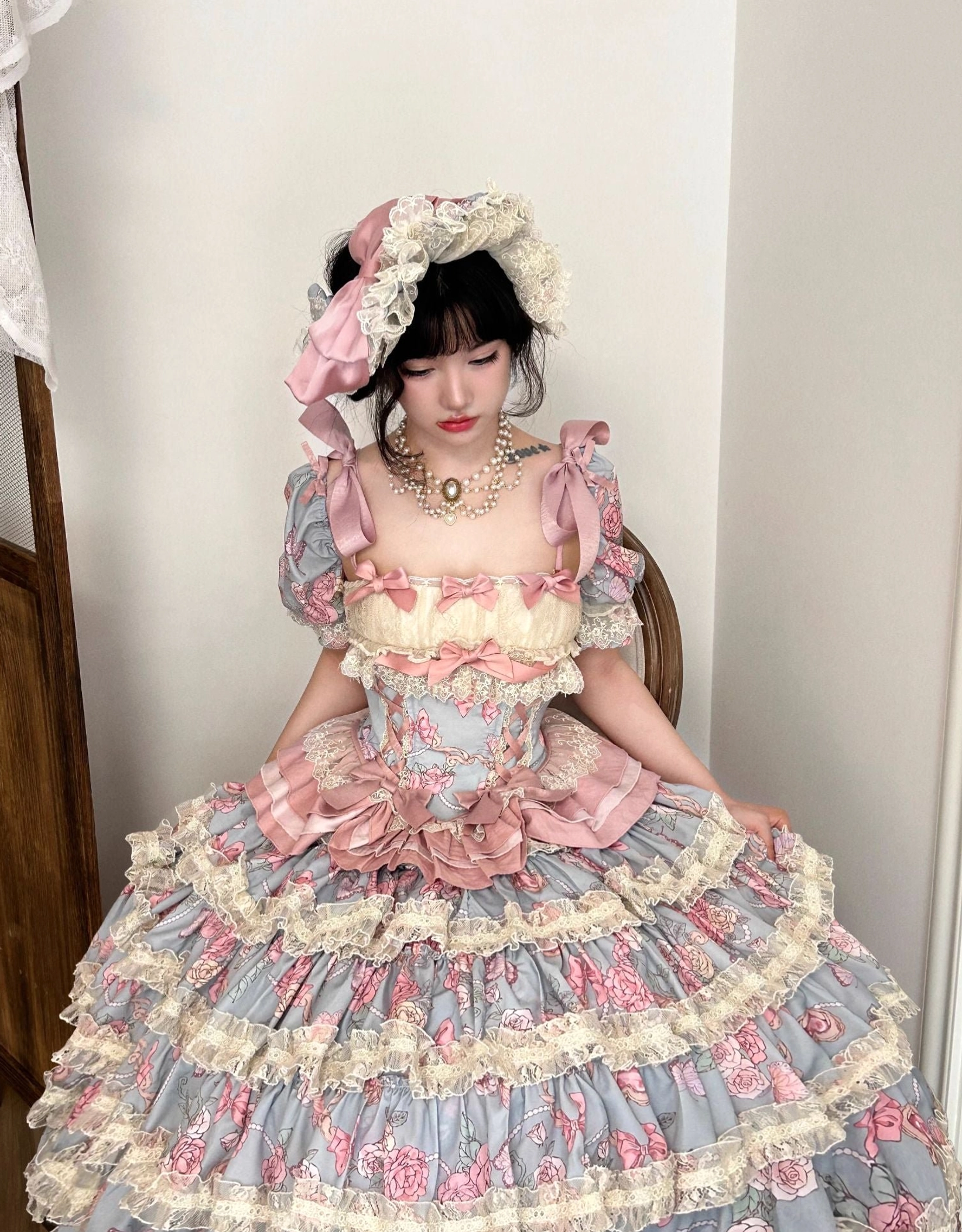 Three-quarter side shot of floor-length sweet lolita dress featuring large front pink satin bow multi-layered ruffle skirt with lace trim and fitted corset bodice worn by model holding skirt to show volume