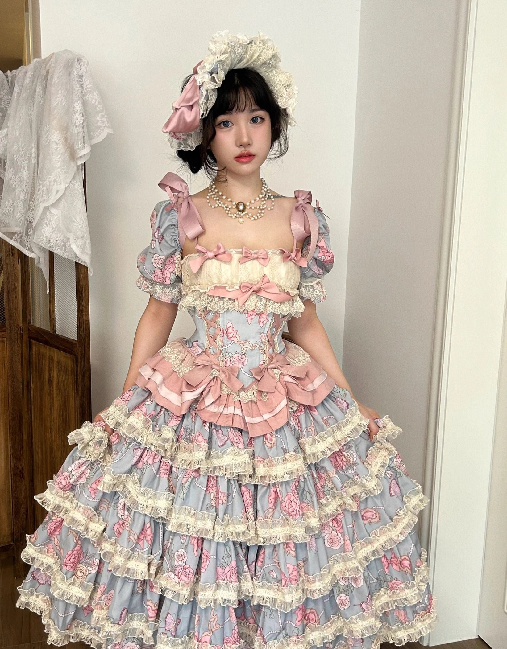 Plus size sweet lolita dress set full front standing shot featuring sky blue base with pink rose floral prints layered ruffle skirt with cream lace edging and puff sleeves with pink satin bows worn with lace bonnet
