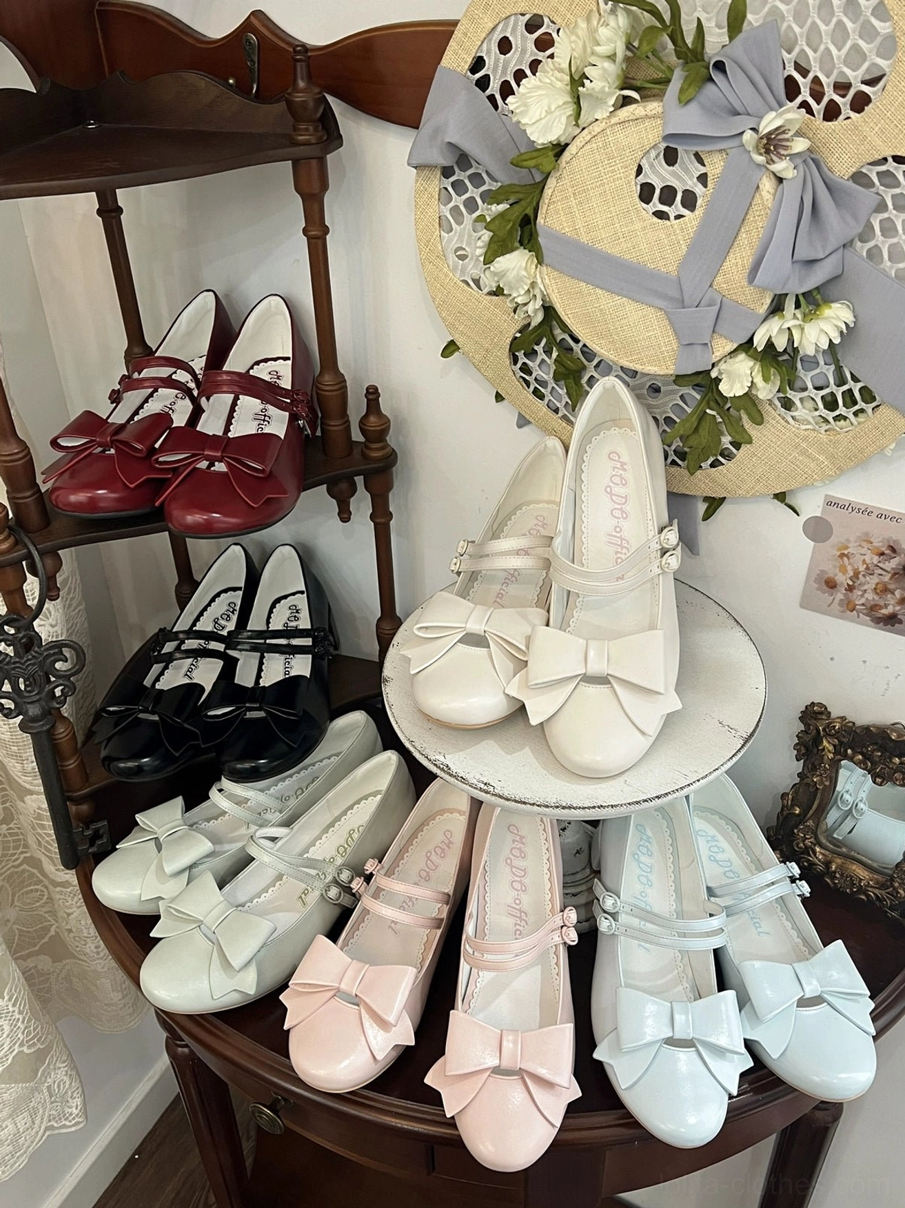 Multi-color pointed toe Lolita Mary Jane shoes in black white pink blue gray displayed overhead on vintage wooden furniture with decorative straw hat and classic bow ankle strap design