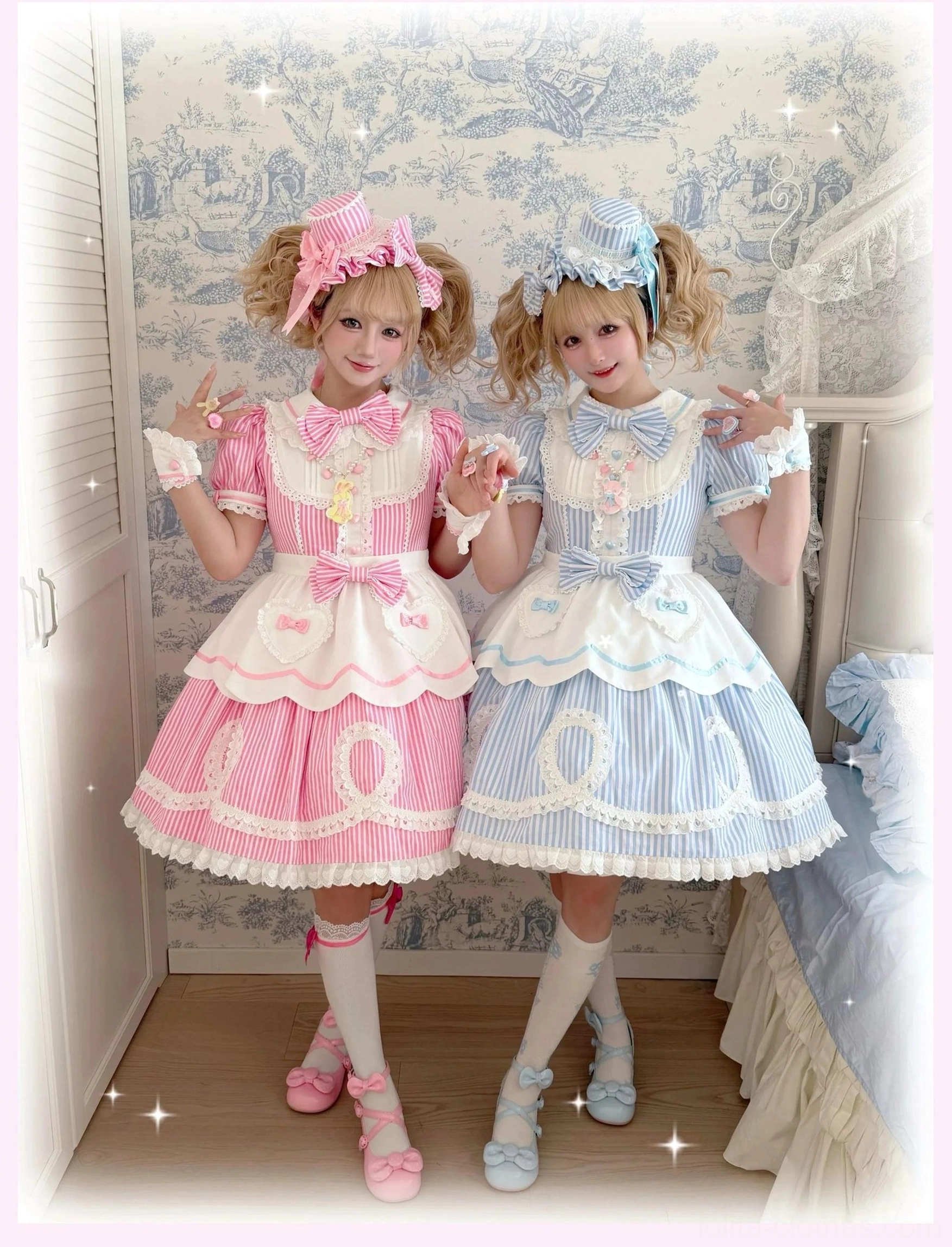 Two posing female models in pink and blue striped lolita OP dresses wearing matching bonnets and coordinated socks with shoes in vintage wallpaper room