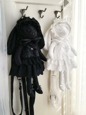 White lamb Yuki and black kitten Nana plush Lolita crossbody bags with lace frill skirts and bow decorations hanging on vintage metal coat hooks, flat lay display of adjustable straps