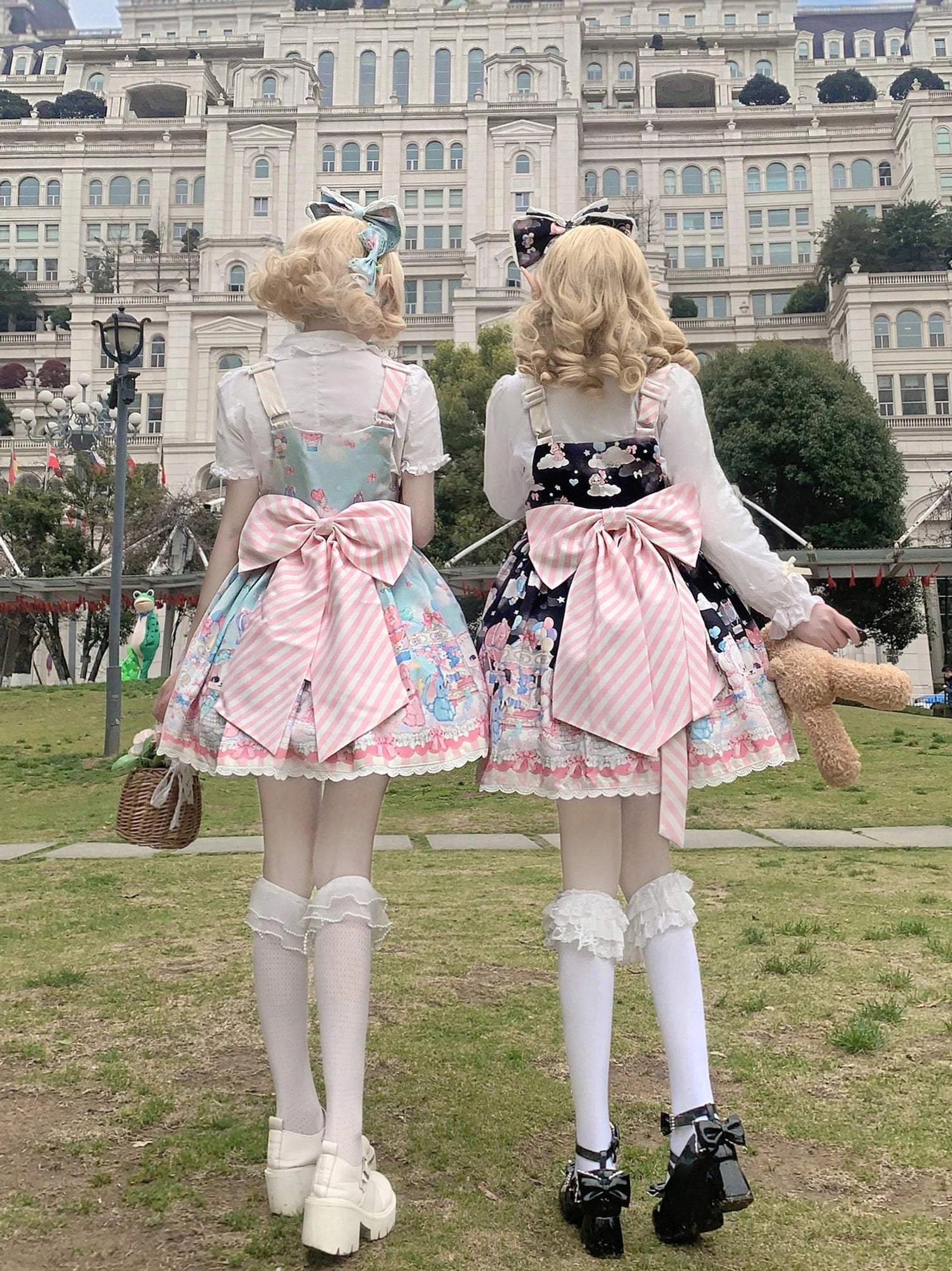 Back full body standing view of two models in puppy themed Lolita salopettes with big pink striped back bows ruffled hem pastel pink and black printed dress versions