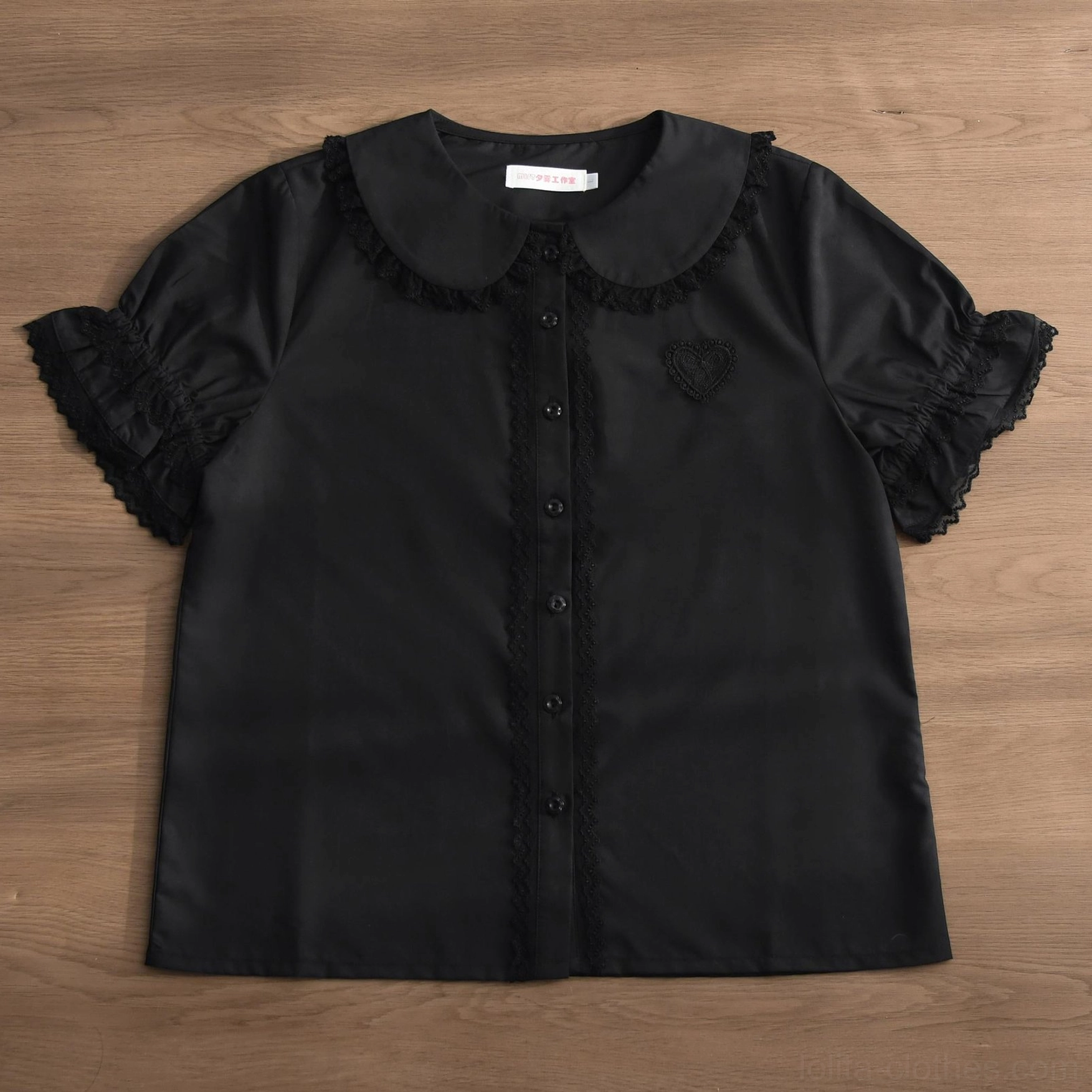Pure black Lolita blouse with lace-trimmed doll collar, ruffled sleeve cuffs and heart embroidery, shown on wooden tabletop