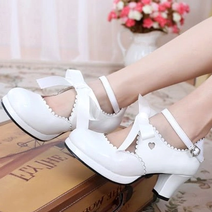 Pure white lolita high heel shoes featuring heart cutout on vamp, scalloped collar, ankle buckle strap, ribbon bow decoration, and smooth white leather displayed on vintage wooden box with flower vase