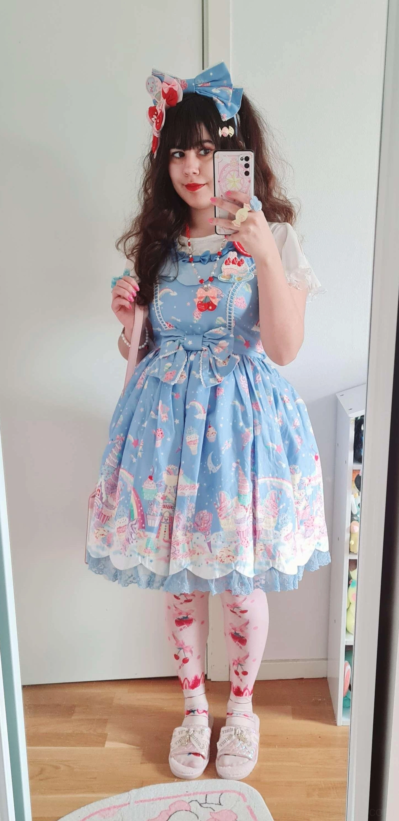 Complete lolita coordinate mirror selfie featuring rainbow gradient petticoat under blue printed dress, matching blue bow hair accessory, printed thigh highs, and white loafers on wooden floor