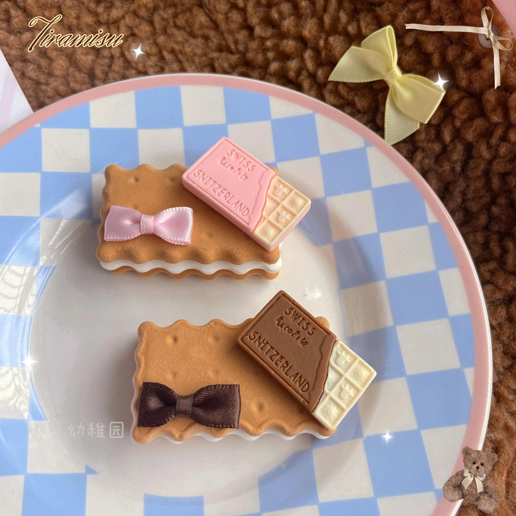 Overhead flat lay of two rectangular cookie lolita hair clips with chocolate bar decorations in pink and brown color variations, featuring white icing edge details on biscuit base and striped bows on blue-white checkered plate