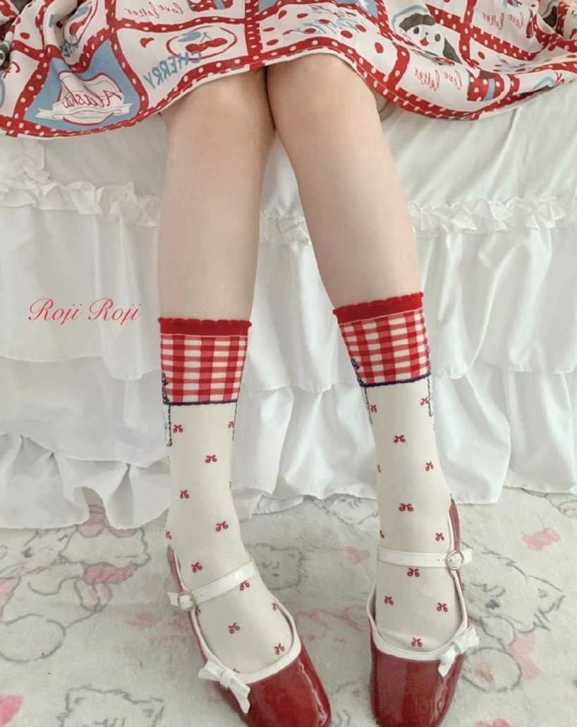 Model wearing red gingham lolita mid-calf socks with cream base and bow prints paired with vintage print dress and red patent leather Mary Jane shoes bedroom setting