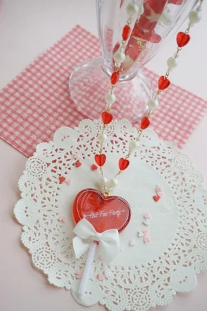Close-up top-down flat lay of red glitter heart lollipop lolita necklace with white fabric bow and beaded chain featuring red heart and pearl beads on gingham background