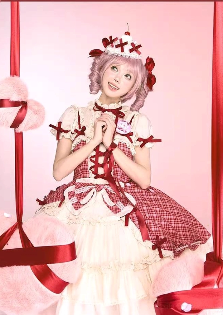 Half-body smiling front view of model wearing red plaid cake lolita JSK dress with cross cake hair accessory, hands clasped under chin, pink ribbon and plush prop background