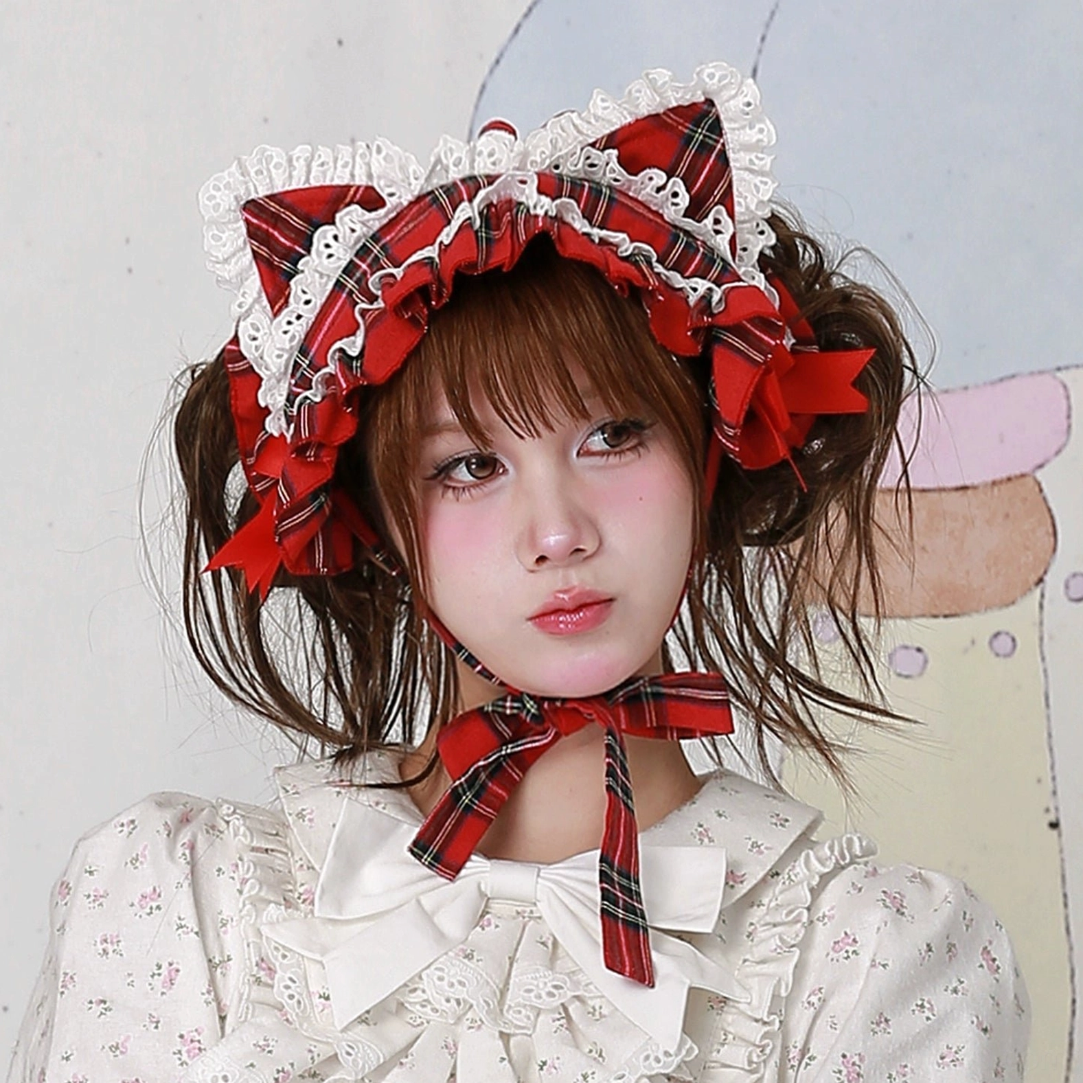Red plaid tartan cat ear hairband with white ruffle lace edging and matching red bow accents, three-quarter left side portrait view, worn with white floral lolita blouse, showing side profile of structured cat ears and coordinated neck ribbon