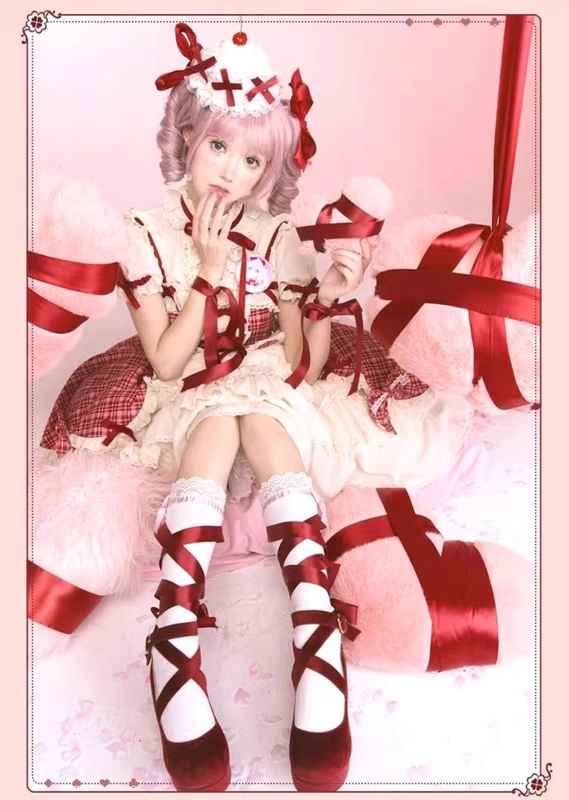 Full-body sitting view of model in red plaid lolita cake JSK dress on pink bedding, showing red strap socks with ribbon bows and Mary Jane shoes, cross cake hair crown