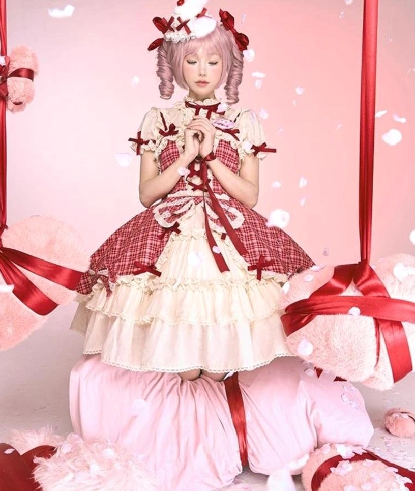 Full-body front standing view of model in red plaid lolita cake JSK dress with hands clasped in front, pink curly twin-tails hairstyle, plush gift box prop background