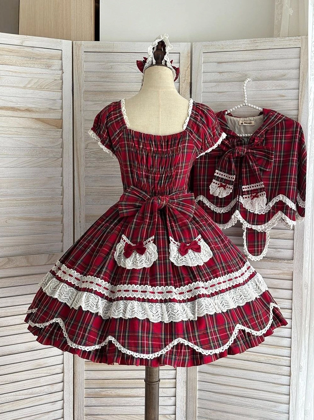 Back view of red tartan plaid lolita OP dress on dress form showing large decorative back bow with heart-shaped lace trim and scalloped hem detail