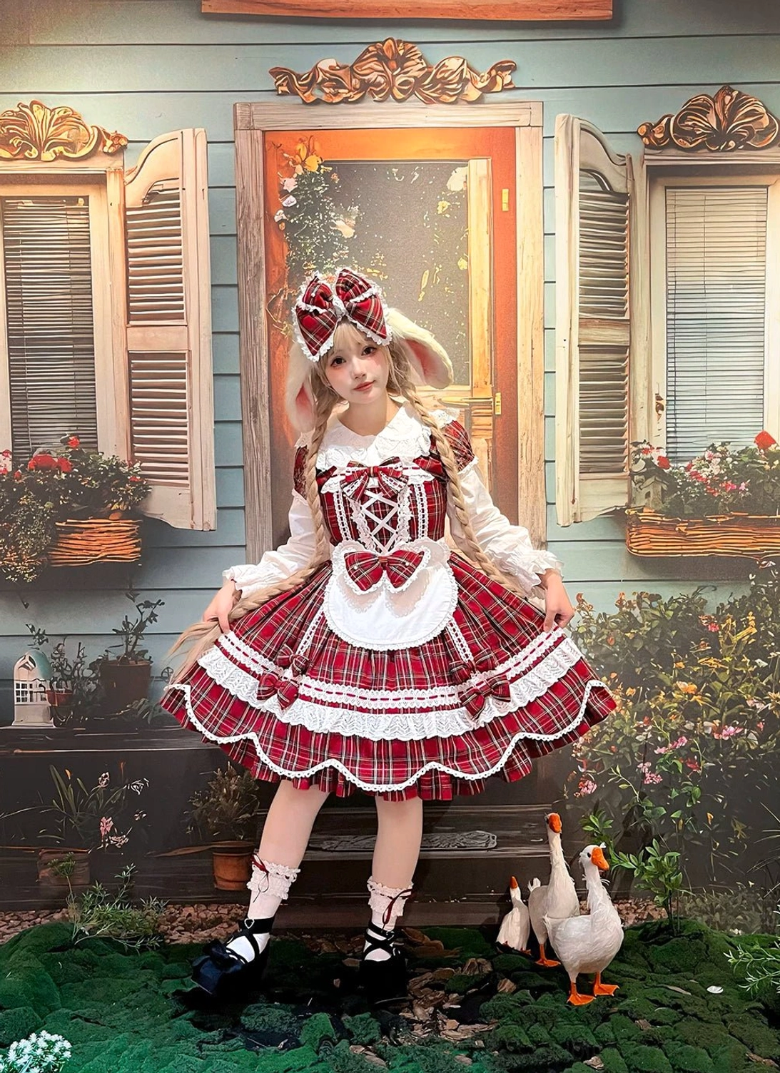 Three-quarter front view of red plaid maid-style lolita OP dress with white apron detail, matching hair bow, black Mary Jane shoes, vintage cottage garden setting