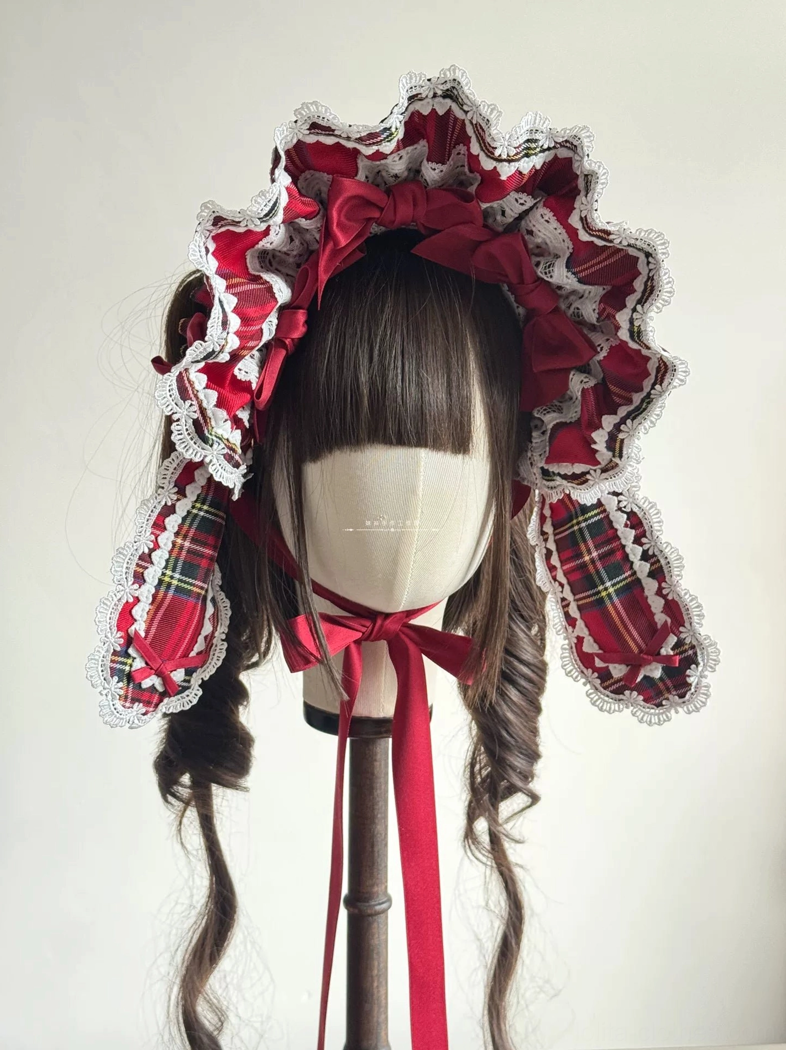 Front three-quarter angle of red tartan Lolita BNT with ruffled lace trim on mannequin head showing ear flap details, white ruffled bonnet brim construction, and wine-red satin bow accent