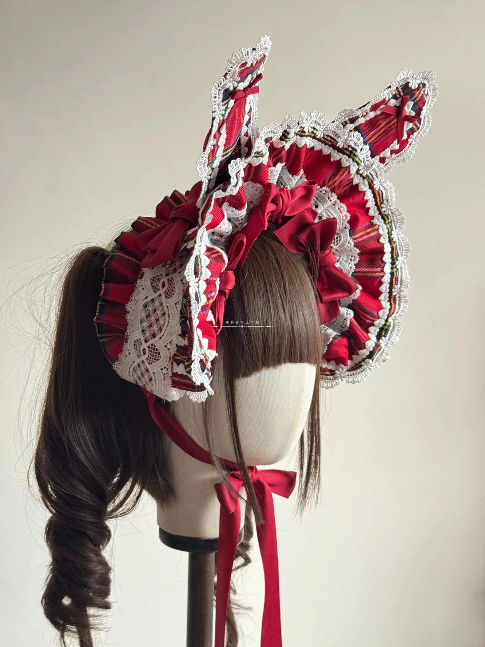 Wire-edged red tartan Lolita BNT bunny ear headdress shown from left rear angle on mannequin with curly brown wig, displaying droopy ear position, white lace trim, and chin tie ribbon detail