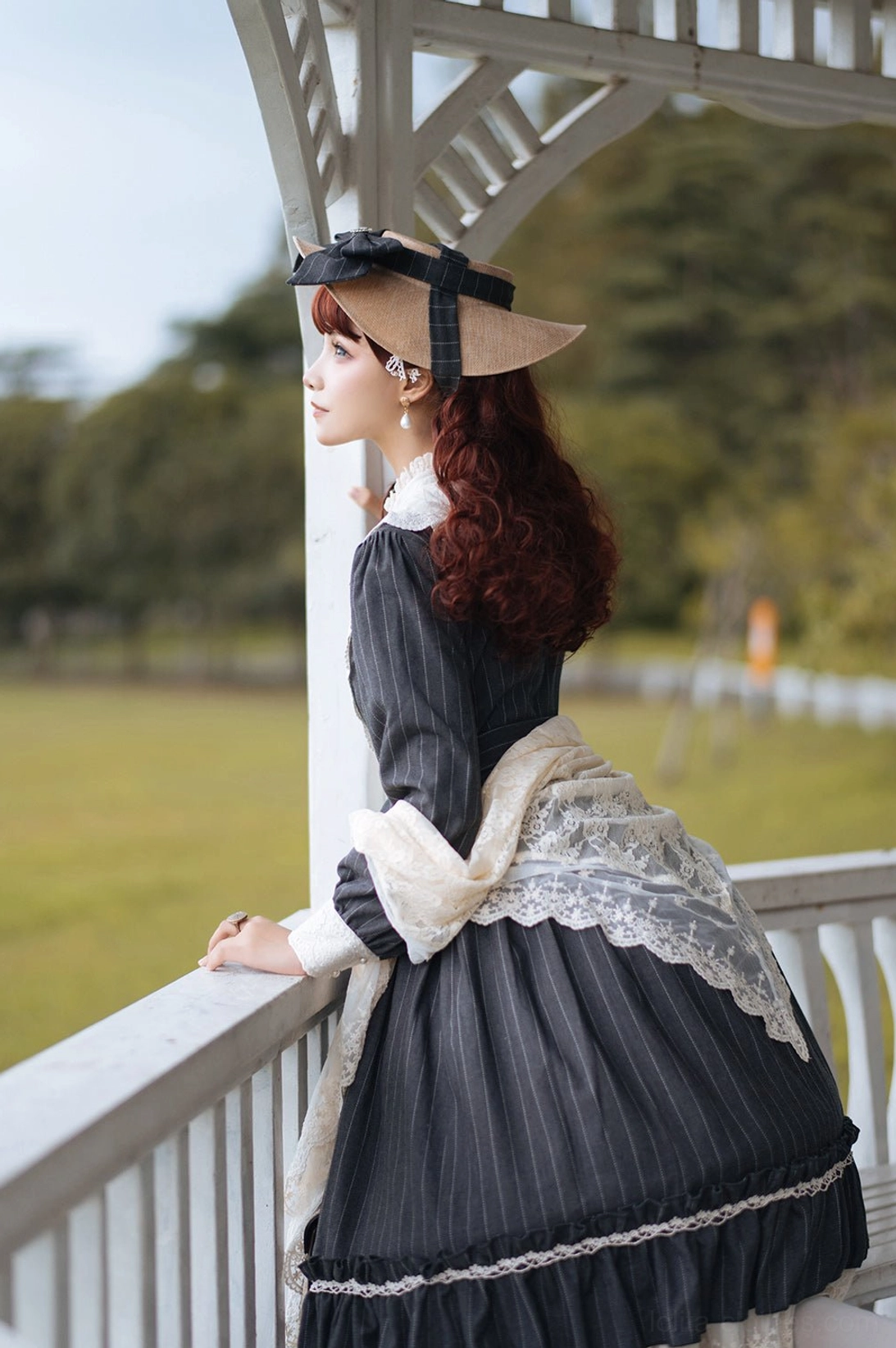 Lolita one-piece dress back view in dark grey striped fabric with large lace sleeve trim, ruffled skirt hem and matching bonnet, standing in garden pavilion balcony setting
