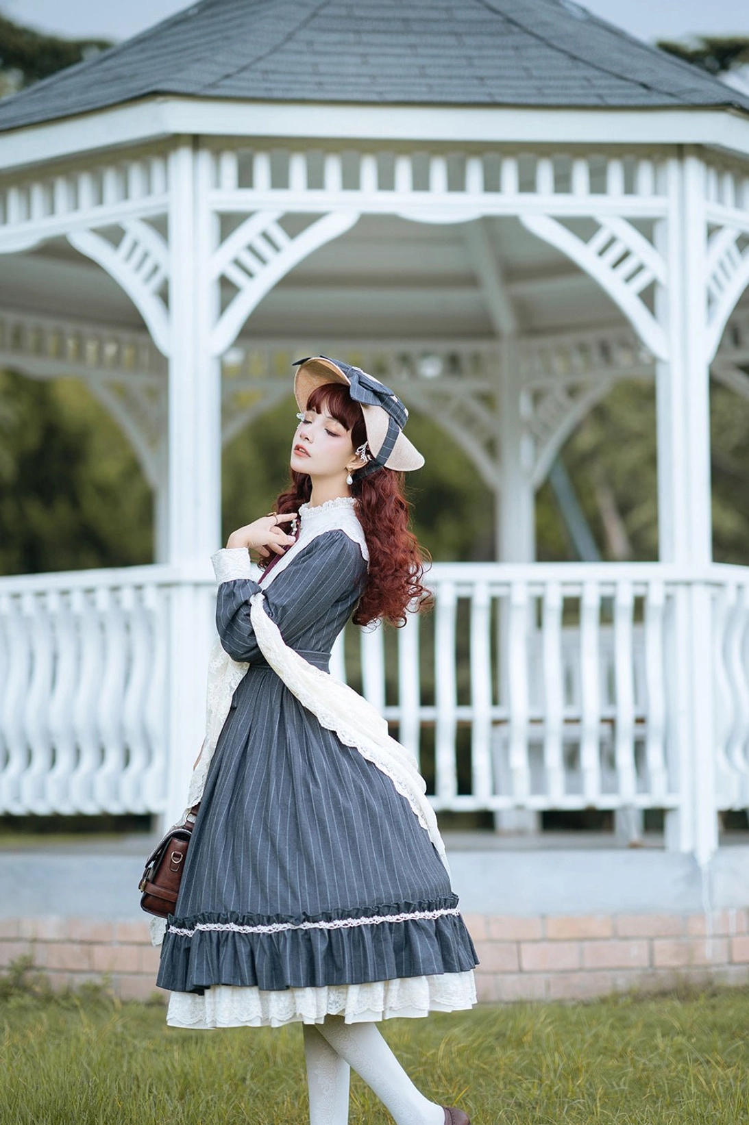 Navy blue striped Vintage Classic Lolita OP dress full back standing portrait with ruffled skirt hem, matching bonnet and brown crossbody leather bag accessory in garden lawn