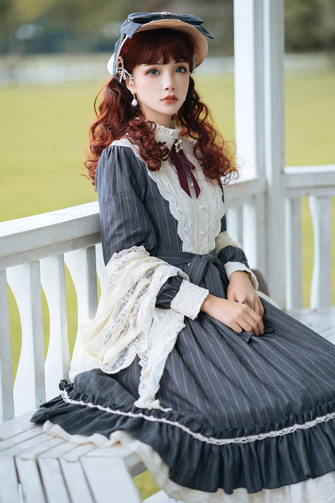 Vintage Classic Lolita one-piece dress with navy blue striped pattern, white lace trims, ruffled skirt hem and burgundy neck bow with matching bonnet, full front sitting on bench