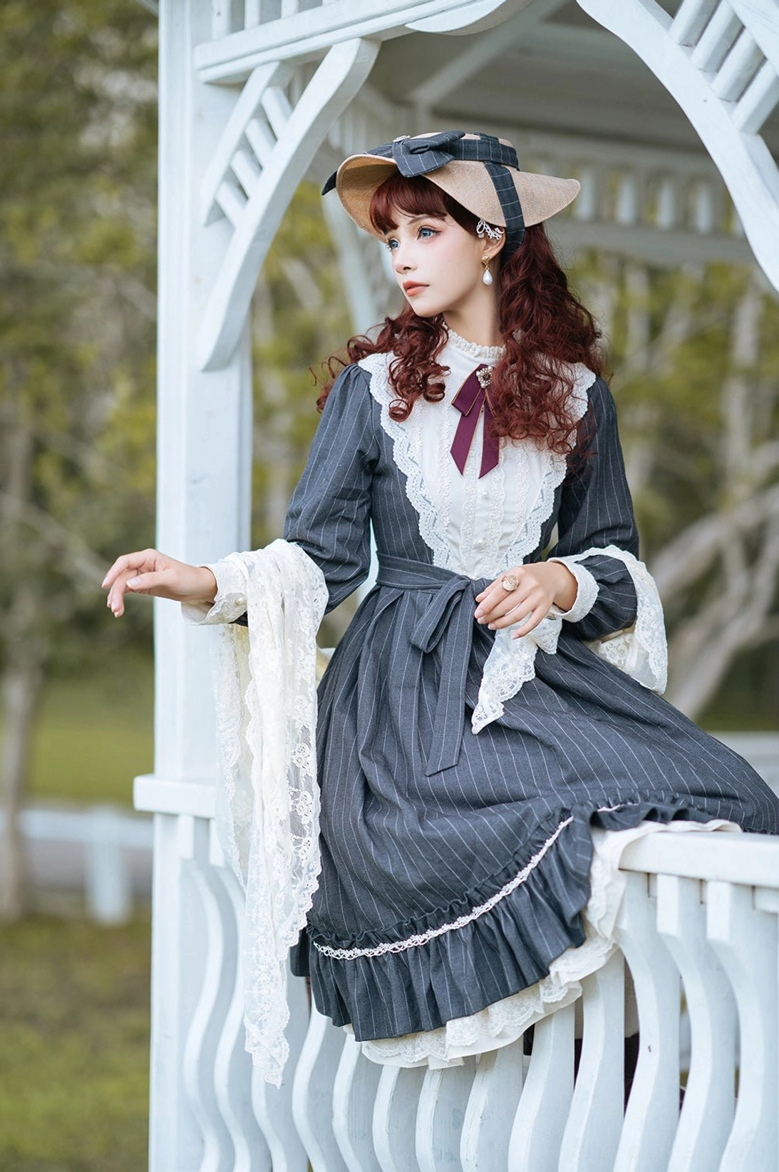 Classic Lolita OP dress featuring navy pinstripe fabric, white lace yoke and ruffled hem with burgundy neck bow and matching bonnet, half-body sitting portrait detail view