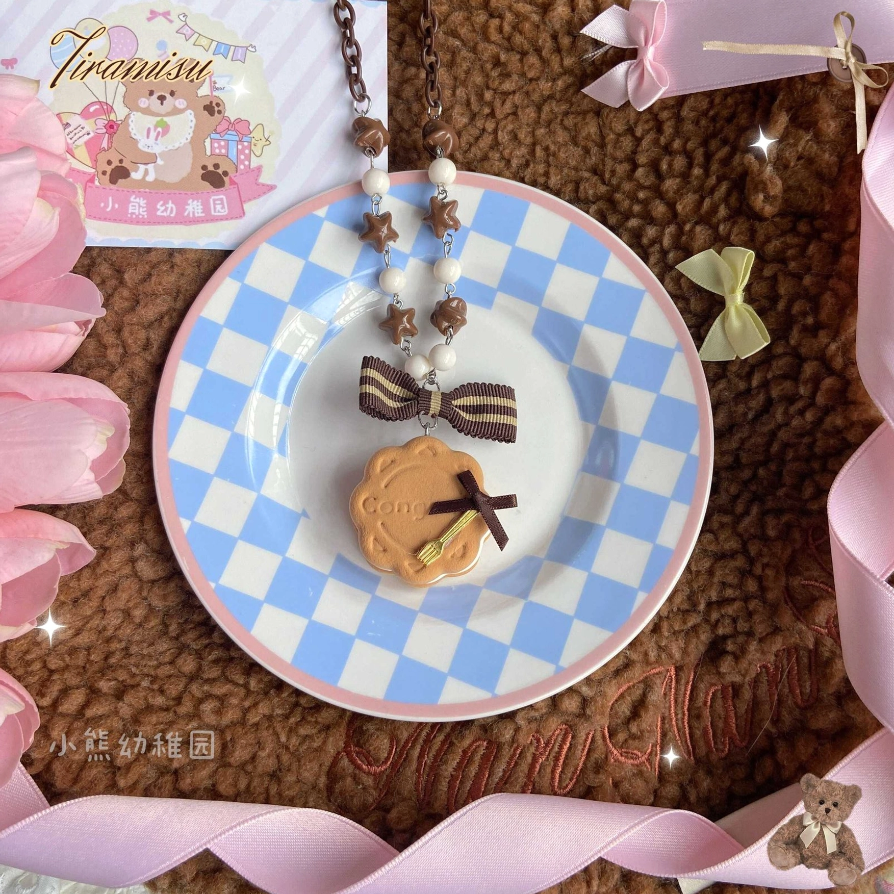 Flat lay close-up of round cookie style lolita hair clip and matching beaded chain with brown bow, tiny fork charm on round biscuit, and striped brown bow chain on pink dessert-themed greeting card background
