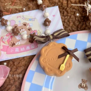 Close-up flat lay of round cookie style lolita hair clip and matching beaded necklace with engraved biscuit charm and tiny fork ornament on blue-white checkered ceramic plate with teddy bear greeting card backdrop