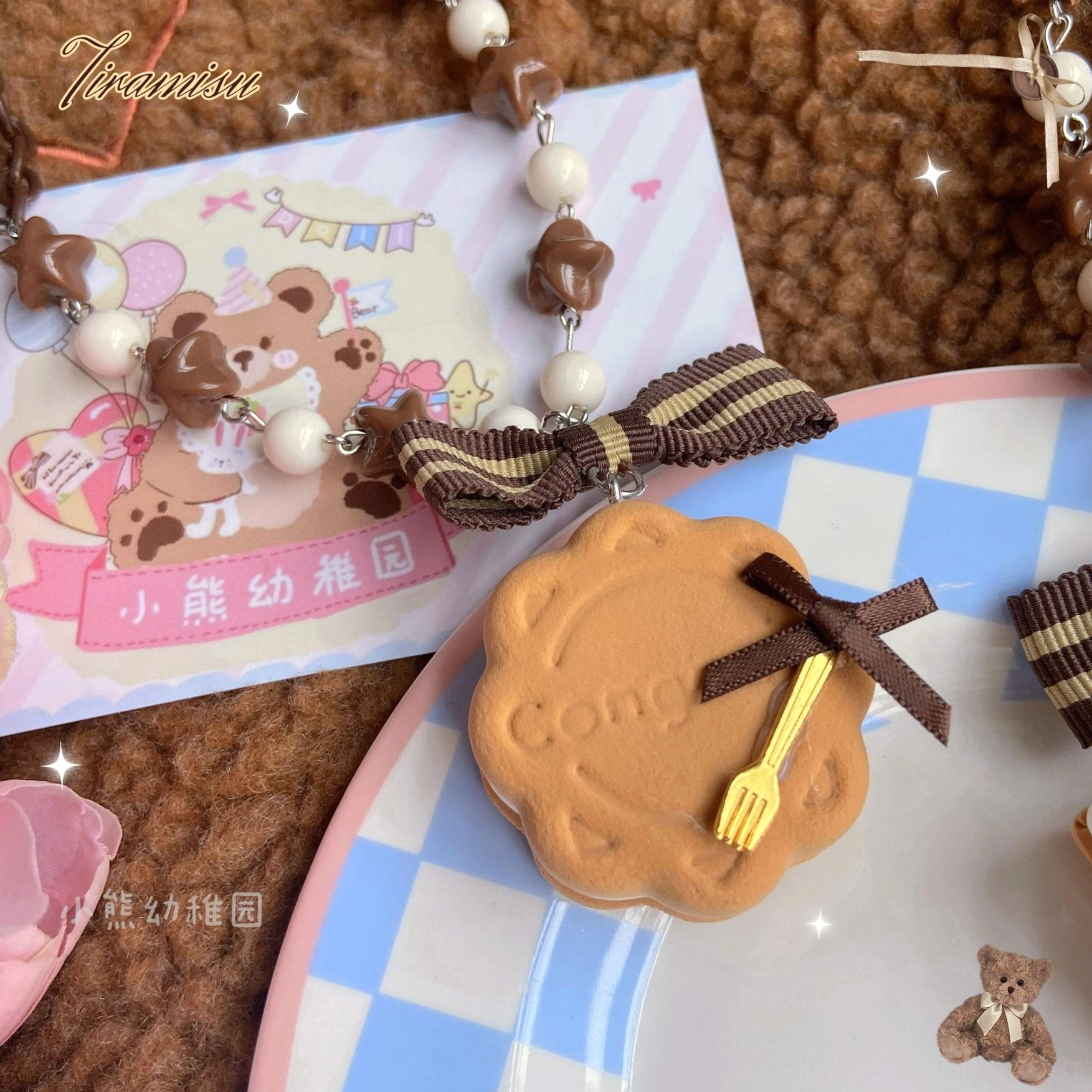 Close-up flat lay of round cookie style lolita hair clip and matching beaded necklace with engraved biscuit charm and tiny fork ornament on blue-white checkered ceramic plate with teddy bear greeting card backdrop
