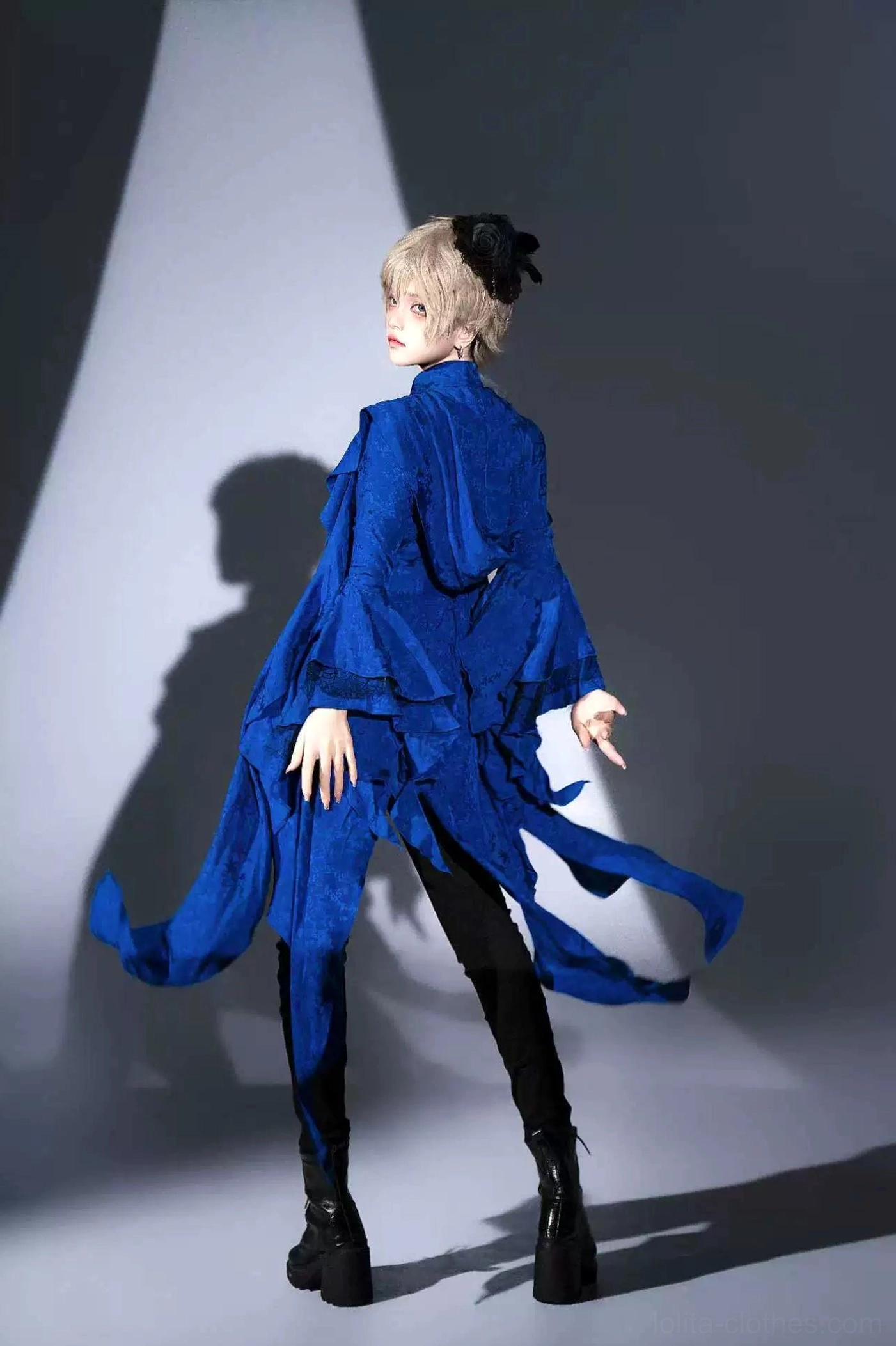 Back view of male model wearing royal blue Ouji Lolita long coat showing trailing asymmetrical irregular hemline, paired with black platform boots and fitted dark pants, studio spotlight backdrop