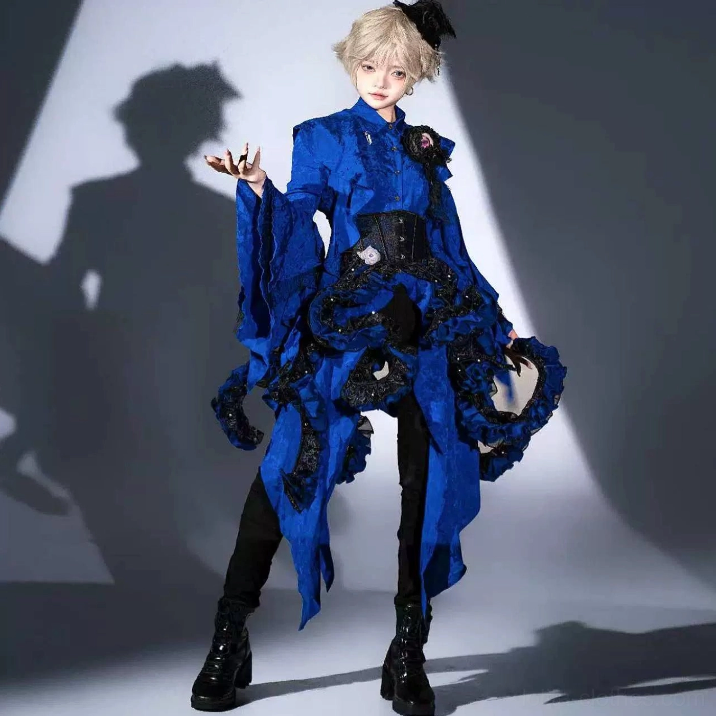 Male model in detailed royal blue Ouji Lolita coordinate set featuring asymmetrical ruffled irregular hem, decorative flower brooch at corset waist, layered flared sleeves, three-quarter front pose