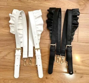 Flat lay of white and black ruffled PU leather lolita bag shoulder straps with gold metal adjustment buckles and secure lobster clasps on wooden tabletop background