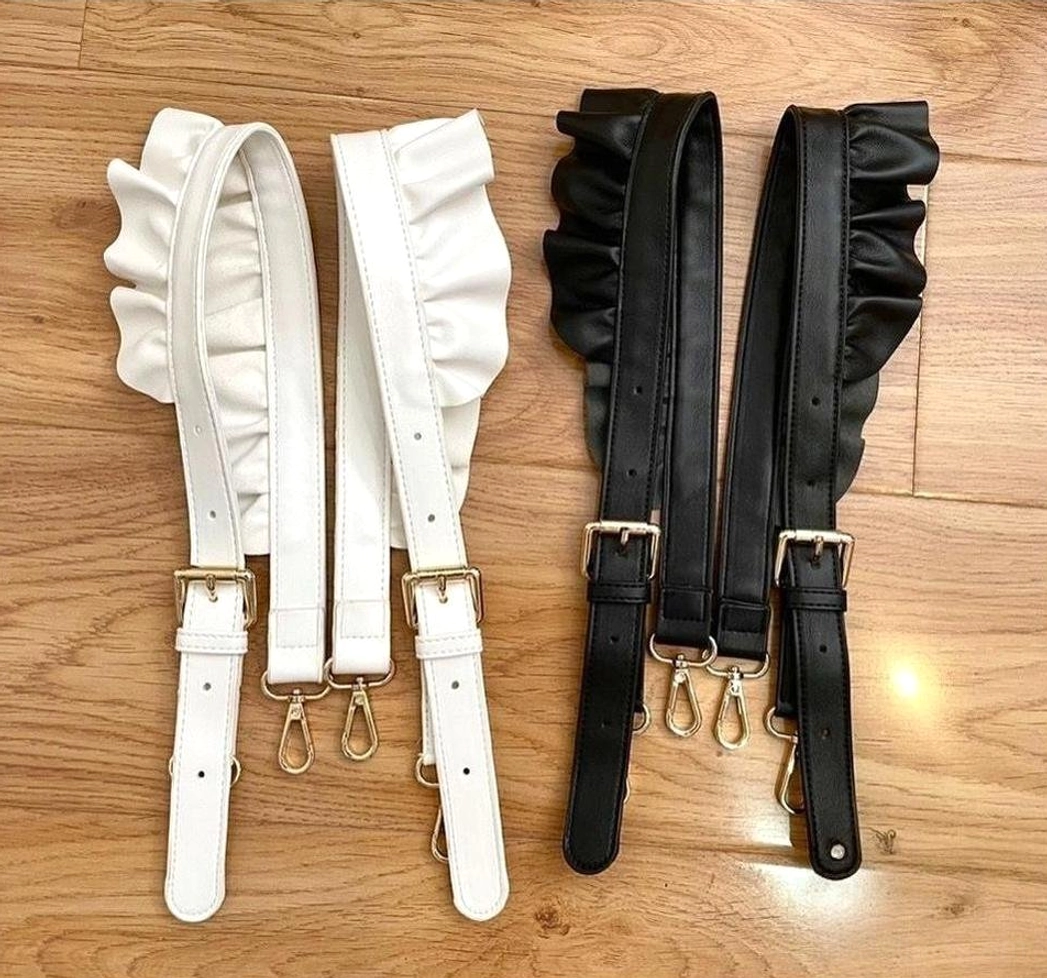 Flat lay of white and black ruffled PU leather lolita bag shoulder straps with gold metal adjustment buckles and secure lobster clasps on wooden tabletop background