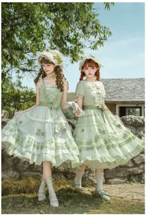 Two models wearing sage green grape embroidered country lolita dresses with tiered ruffles and matching flower bonnets standing on a grassy stone yard full front view