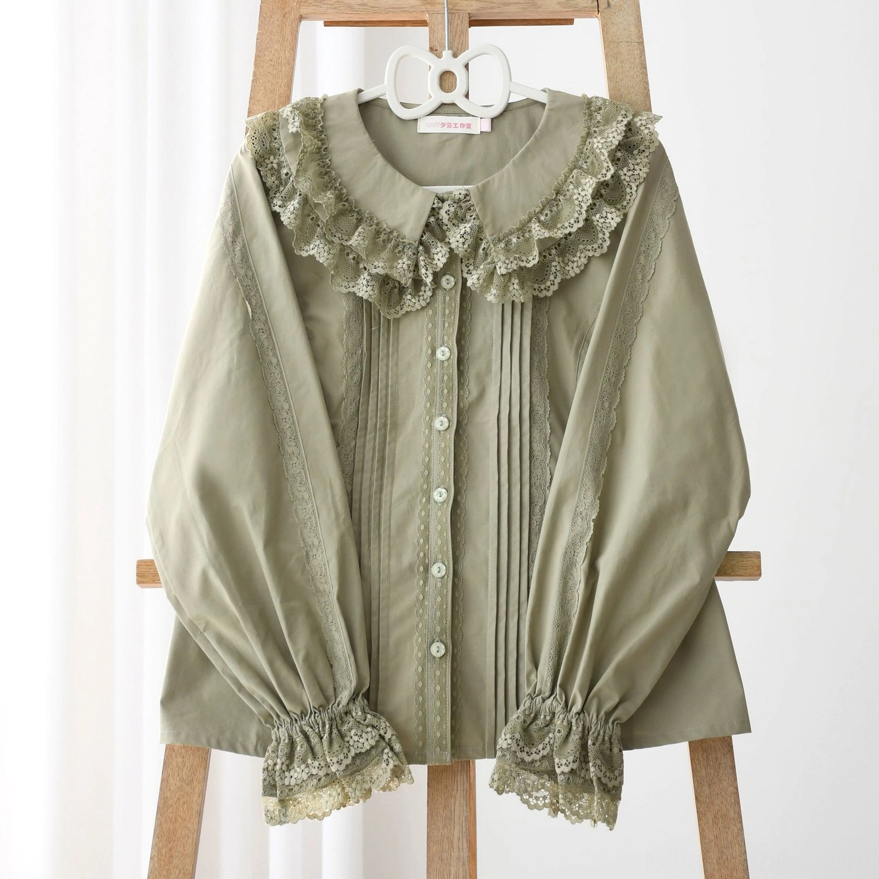 Close-up front flat display of solid sage green Lolita long-sleeve blouse on wooden hanger with white background, showcasing lace ruffled Peter Pan collar, front pleated details, button placket, and lantern sleeves