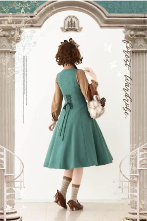 Sage green Lolita jumper skirt with lace-up adjustable back waist closure, back full-body view displaying midi flared skirt silhouette paired with tan inner blouse, model styling with plush handbag and brown loafers