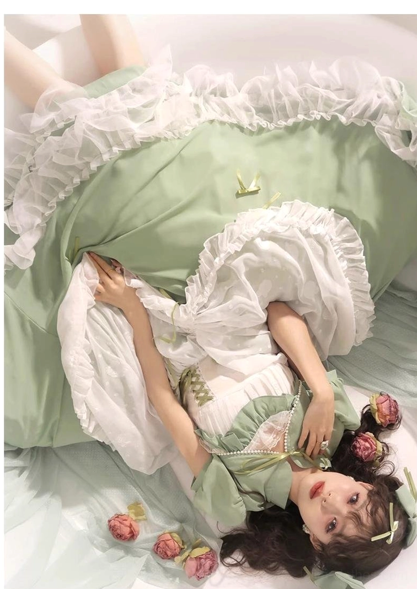Overhead lying-down view of sage green plus size wedding lolita JSK on green bedding, showcasing ruffled petticoat, voluminous skirt, golden bow details, and full silhouette