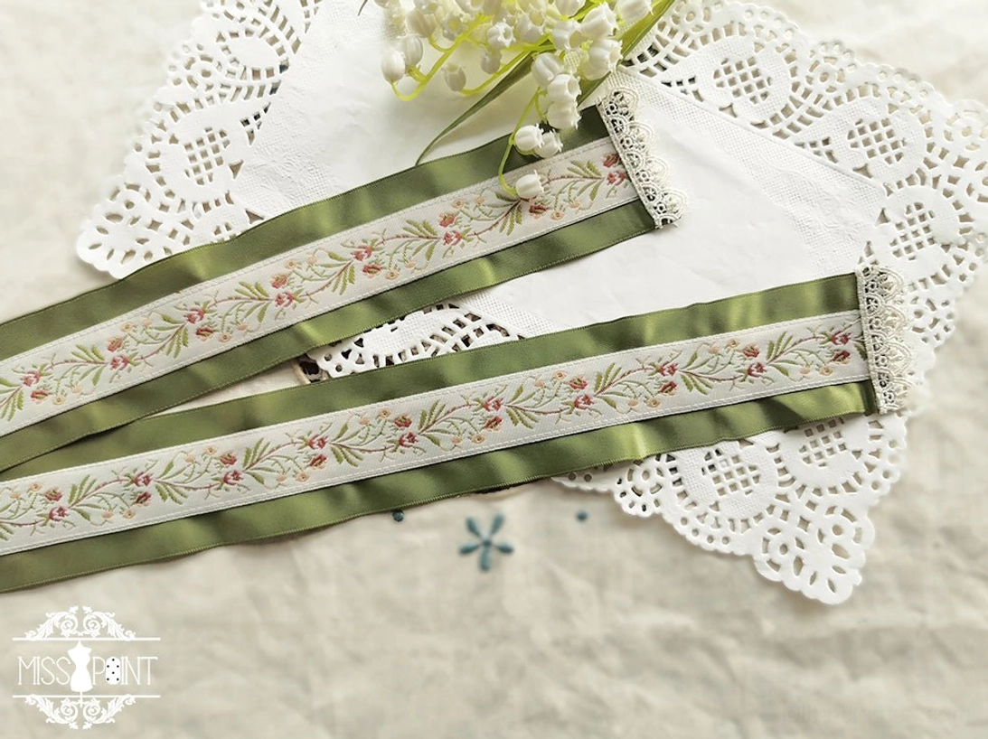 Flat lay overhead view of sage green embroidered lolita waist ribbon belt with delicate floral weaving and pearl decorations on lace doily
